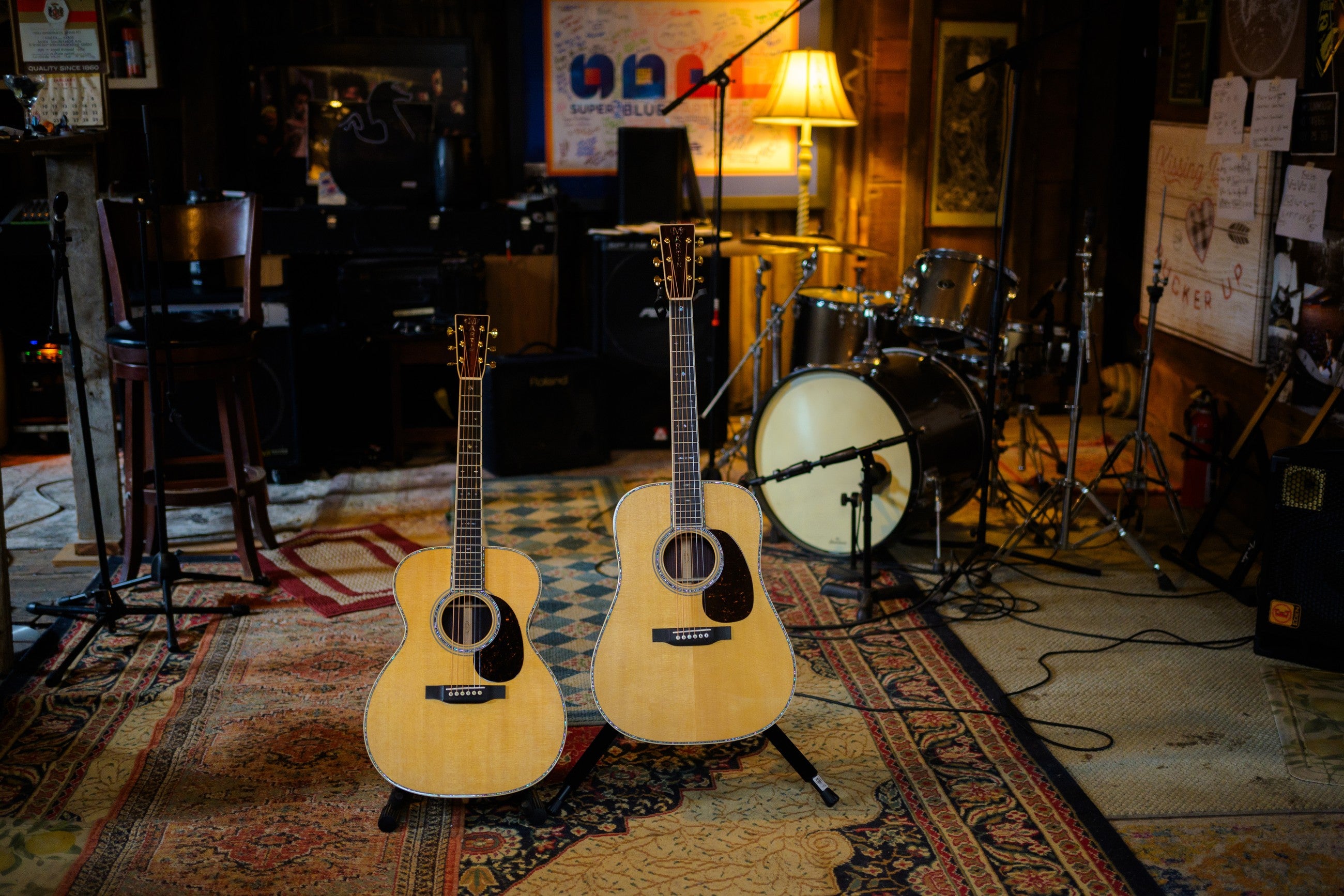 Two Martin acoustics standing on a Persian rug in a warm studio with amps and a drum kit. Setups and repairs at Paul's Guitar Hideout.